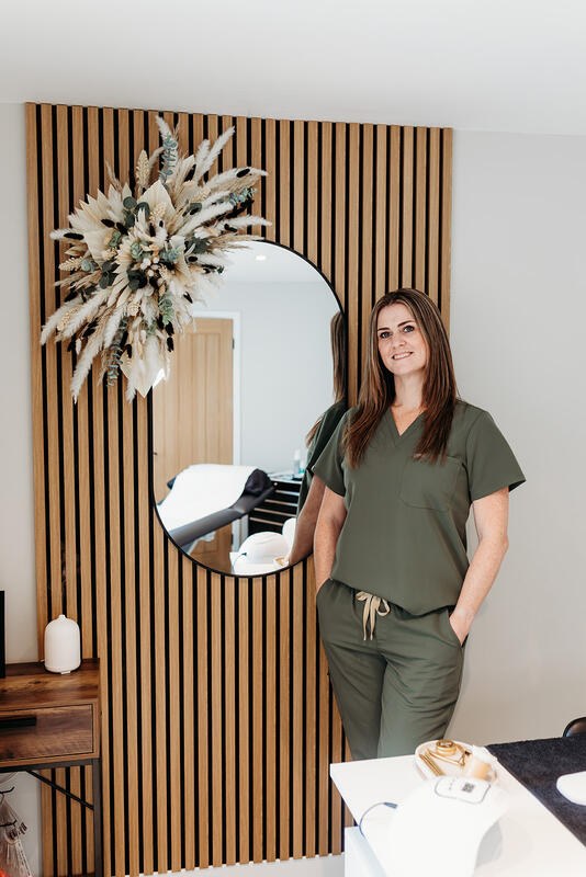 Vicky beside the slatted oak wall and mirror in her treatment room